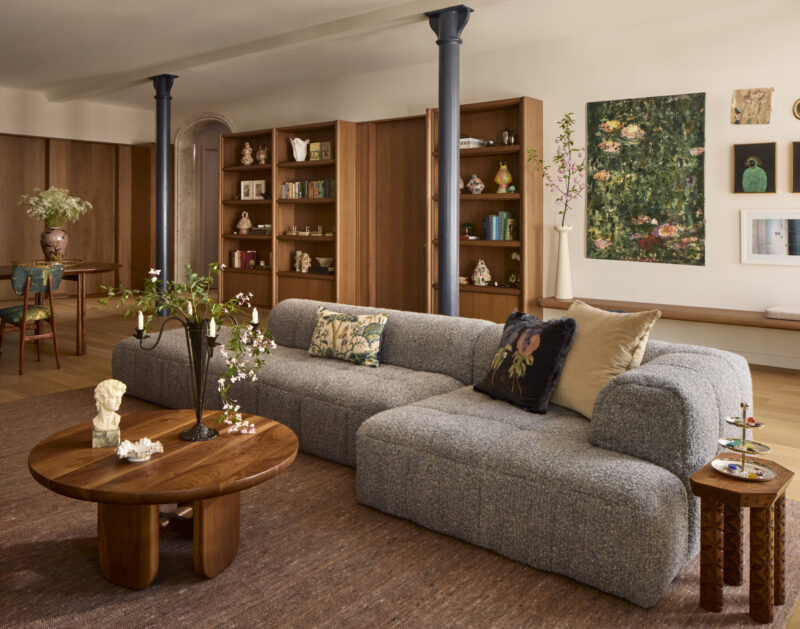 A modern living room with a grey sectional sofa, wooden coffee table, bookshelves, artwork on the walls, and decorative plants.