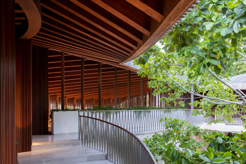 A modern building corridor with wooden beams, metal railings, and greenery from nearby trees, featuring natural light and an open, airy design.