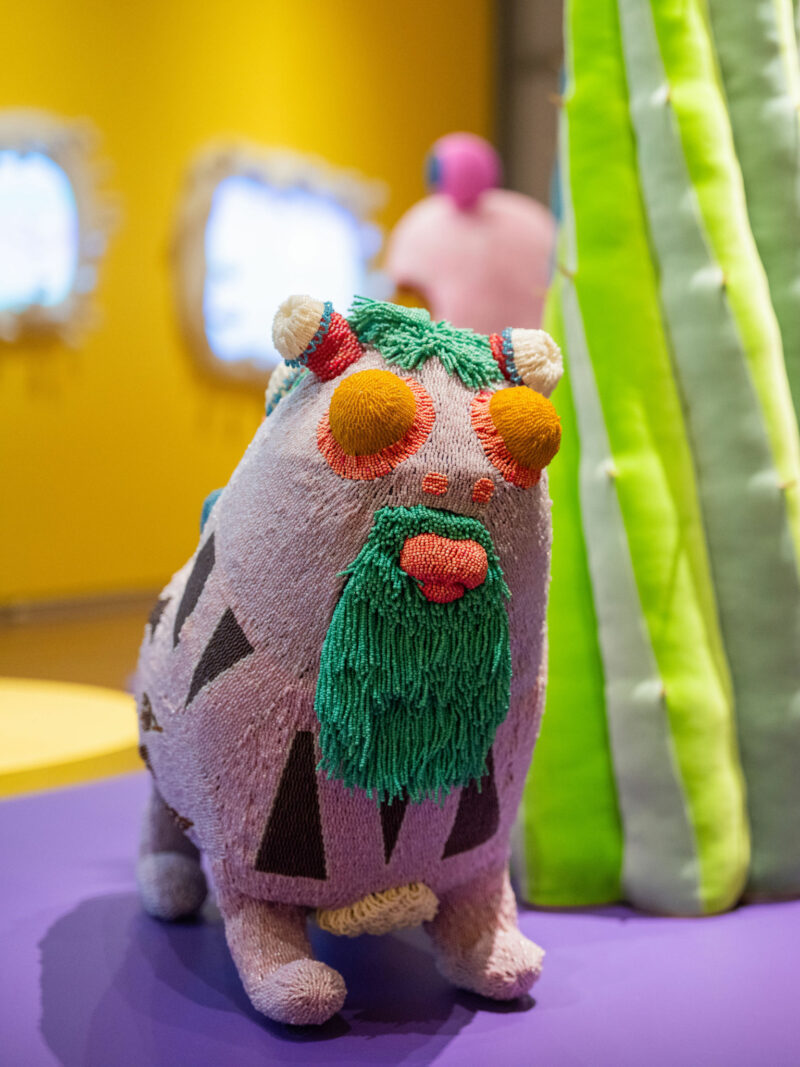 A colorful, yarn-covered animal sculpture with a green beard and orange eyes stands on a purple surface near a green, cactus-like structure, evoking the whimsical style of Haas Brothers Uncanny Valley.