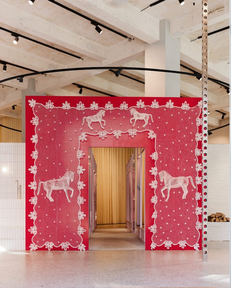 A decorative pink and red wall with white horse and floral designs frames an entryway inside Baum und Pferdgarten store with exposed ceilings.