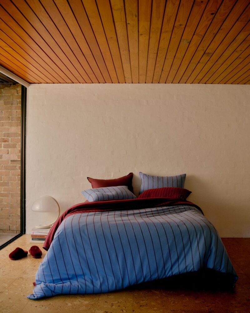 A neatly made bed with blue and maroon striped bedding from Hommey, pillows, and slippers against a white brick wall under a wooden ceiling. A lamp and books are on the floor nearby.