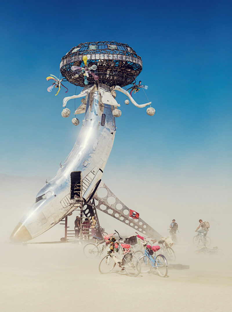 A large art installation resembling a futuristic aircraft nose and metal structure rises from a sandy desert, with people on bicycles and a blue sky in the background.