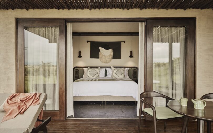 A cozy bedroom with a large bed, decorative pillows, wall art, and pendant lights, viewed through open sliding glass doors from a patio with chairs and a small table. Designed by Luxury Frontiers.