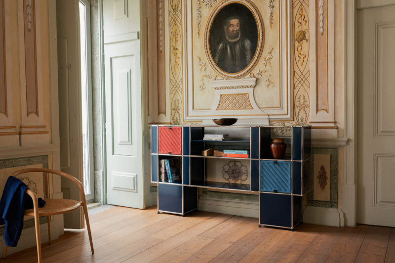 Open doorway in ornate room with wooden floor, blue modular shelving unit from USM holding books and decor, chair to the left, and a portrait of Armando Cabral above the shelves on a decorated wall.