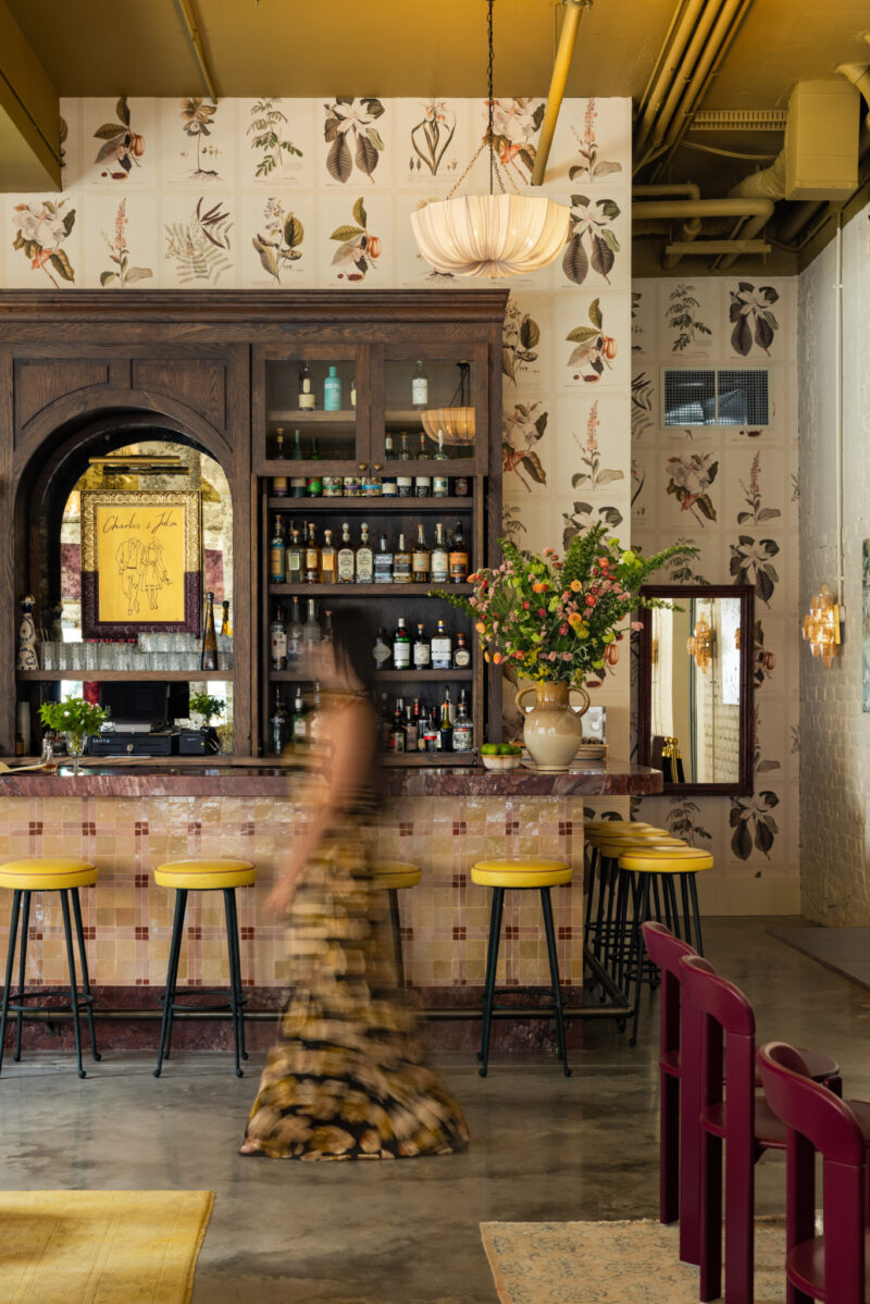 A woman in a floral dress walks past the Charles & Julia bar with yellow stools, a large floral arrangement, and shelves of bottles, in a space decorated with botanical wallpaper.