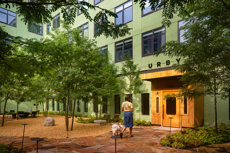 A person stands with a dog in a landscaped courtyard outside Fishtown Urby, a green apartment building with a wooden entrance labeled "URBY.