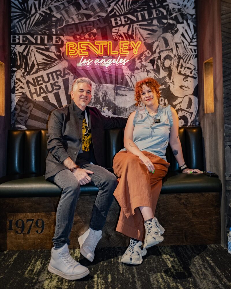 Two people sit on a black bench under a neon "Bentley Mills Los Angeles" sign, with a comic-style collage wall creating an iconic landmark vibe in the background. Land/Mark