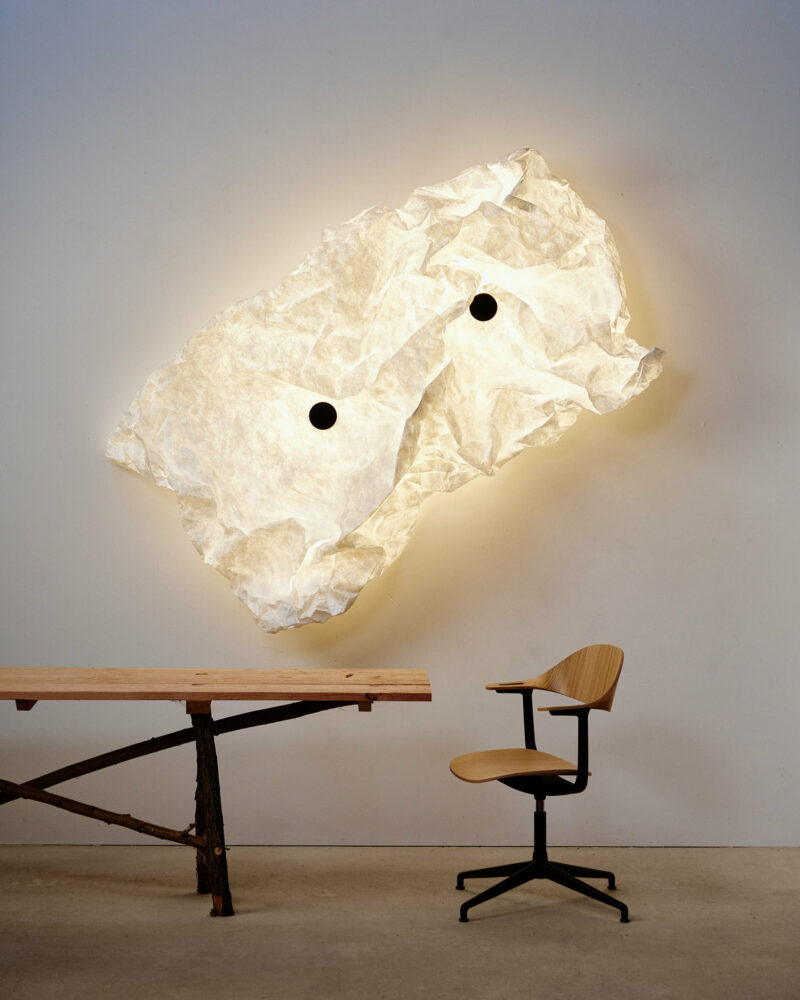 A sculptural light fixture resembling crumpled paper is mounted on a white wall above a black and wood desk and a wooden swivel chair.