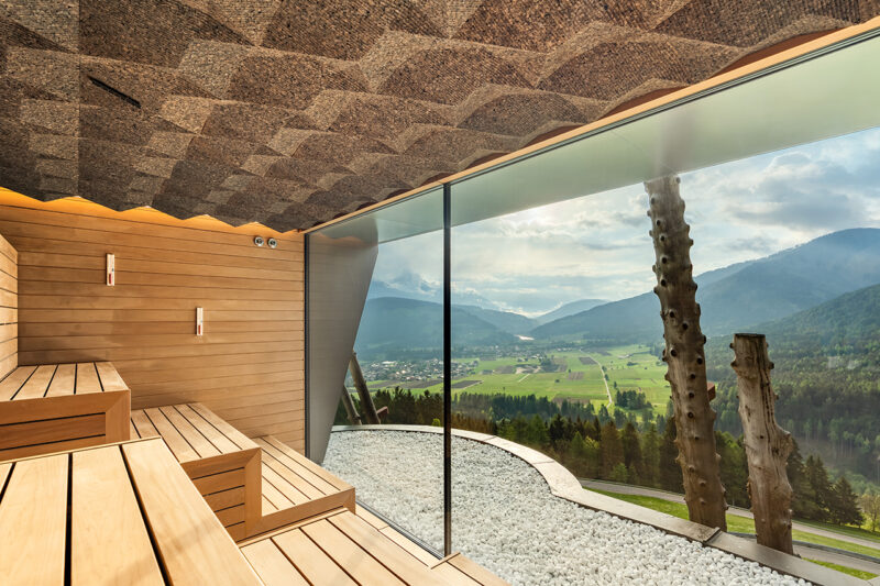Modern wooden sauna with tiered seating, large floor-to-ceiling window offering a panoramic view of a green valley, mountains, and cloudy sky. White stones decorate the floor near the window, reflecting the refined design seen in the Hotel Hubertus spa expansion.