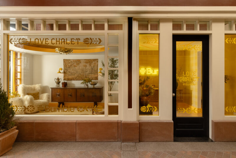 Exterior view of a storefront with yellow-tinted windows, displaying the words "LOVE CHALET" and "Bumble." Inside, cozy furniture and wall art are visible through the glass, reflecting elements of Studio Coco Greenblum’s inviting design aesthetic.