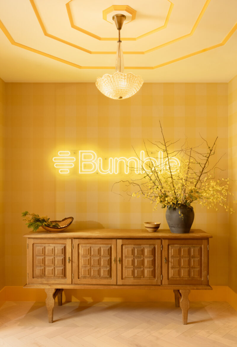 A wooden sideboard with vases and bowls stands below a neon Bumble sign on a yellow plaid wall, under a chandelier in the inviting style of Studio Coco Greenblum.