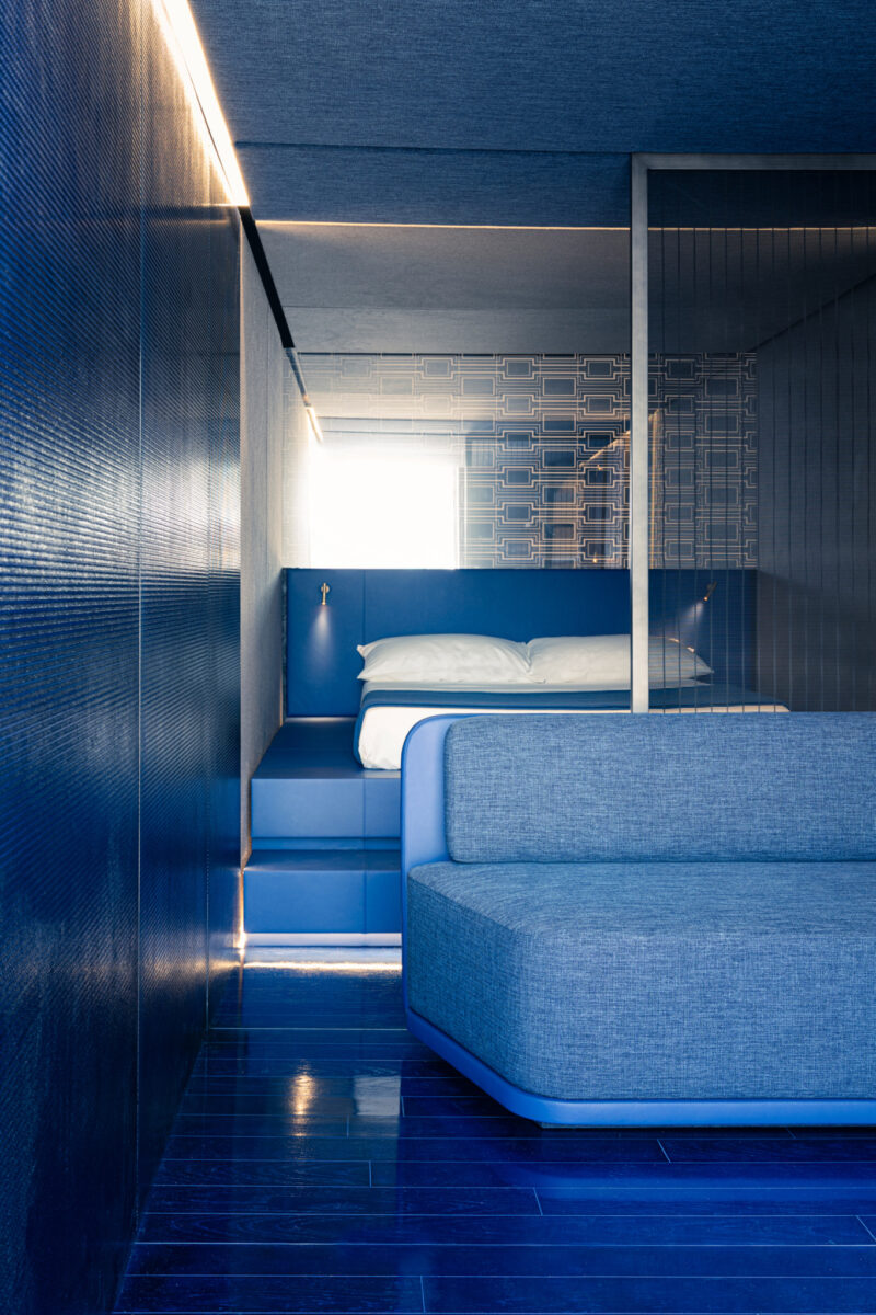 Modern bedroom with blue walls, floor, and furniture, featuring a bed with white linens and a large blue sofa in the foreground—an inviting retreat inspired by the m2atelier Crafted Capsule and illuminated by natural and accent lighting.