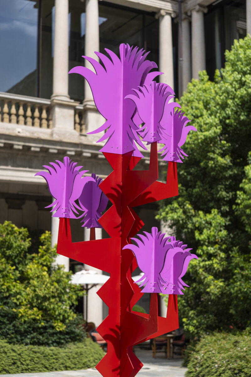 A modern outdoor sculpture, titled Arcipelago Botanico, features a striking red zigzag base topped with several magenta abstract flower shapes. Set in a courtyard adorned with green bushes and framed by a stone building in the background, this artwork seamlessly blends vibrant design with botanical inspiration.