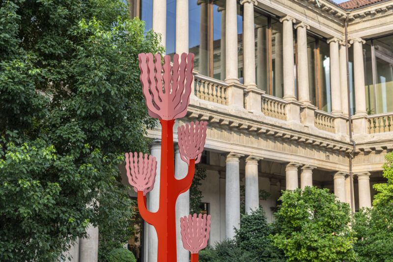 A red sculpture with pink hand-shaped elements stands in front of a classical building with columns, surrounded by green trees, evoking the creative spirit of Arcipelago Botanico.