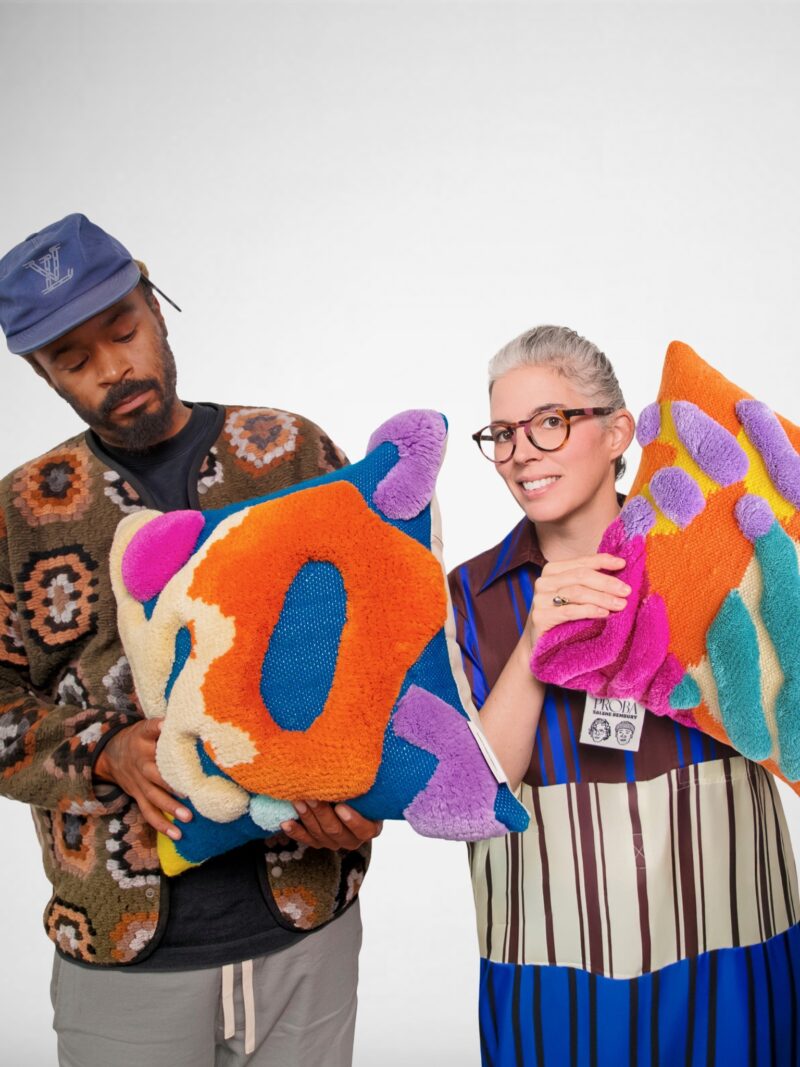 Two people hold colorful, abstract-patterned pillows from the StudioPROBA x Salehe Bembury Soft Imprints collection. The man on the left wears a blue cap and patterned sweater, while the woman on the right wears glasses and a multicolored dress.