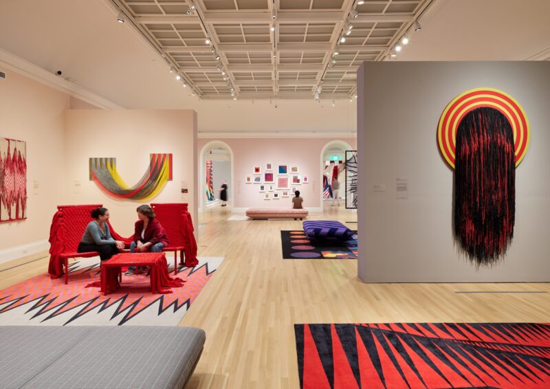 A modern art gallery features vibrant textile artworks, geometric rugs, and two people seated on red woven chairs, engaged in conversation.