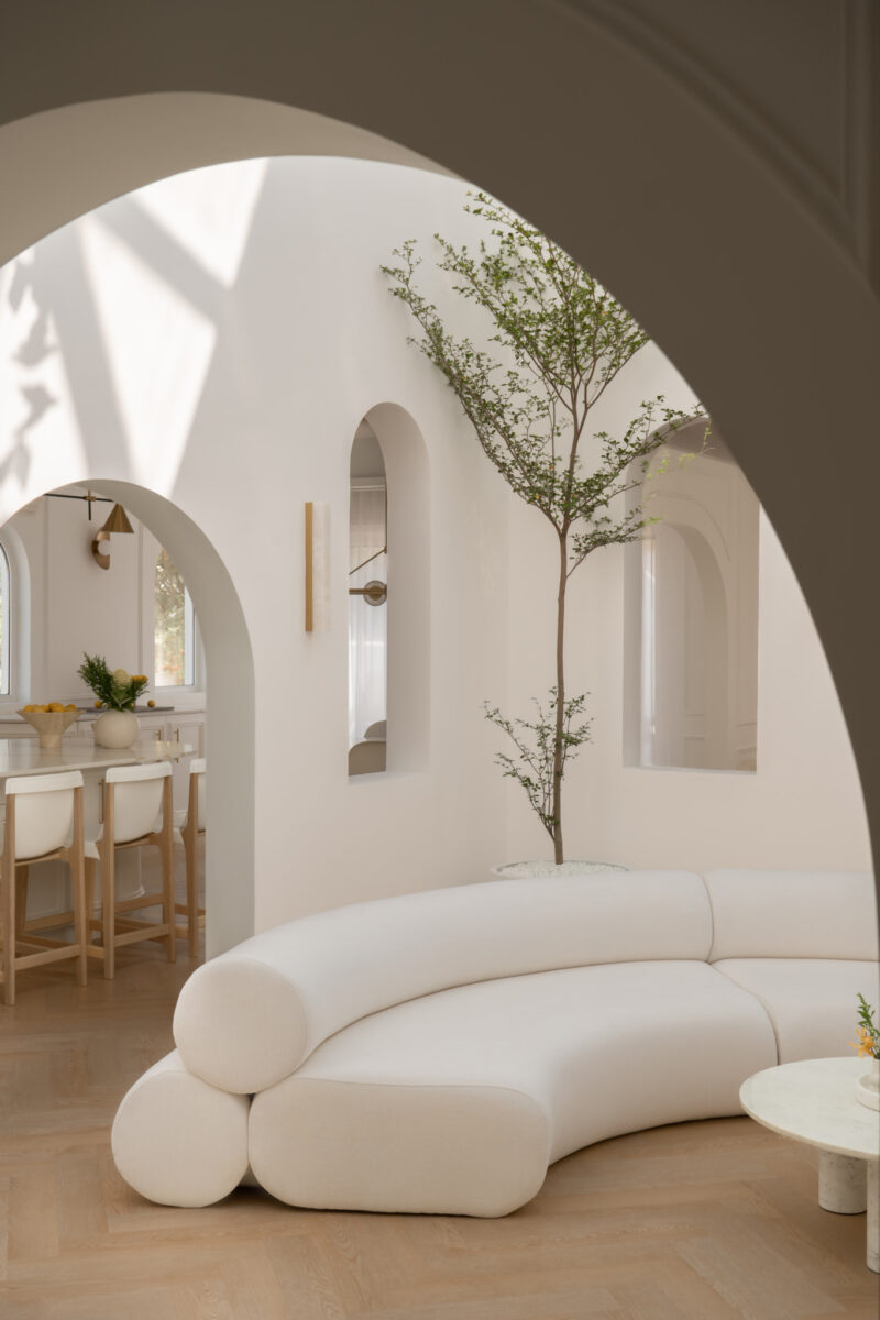 Minimalist living room with curved white sofa, a round coffee table, arched doorways, indoor tree, and light wood floor. Sunlight casts shadows on white walls.