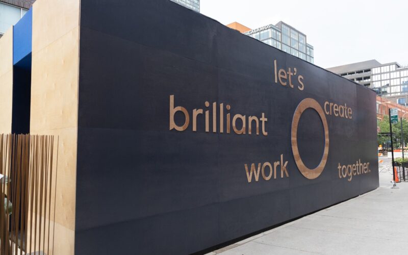 A large black wall with tan text reads: "let's create brilliant work together." This inspiring message, reminiscent of the collaborative spirit at Coalesse Co.Lab, is displayed beside a sidewalk in an urban area.