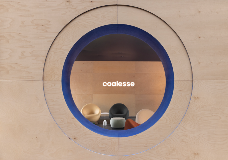 Circular window with a blue border shows a modern seating area with four chairs and a small table, set within the inviting ambiance of the Coalesse Co.Lab, with the word "coalesse" illuminated on the wall inside.
