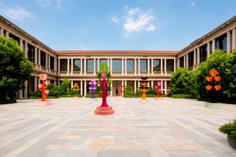 Open courtyard with colorful abstract sculptures from the Arcipelago Botanico collection and a symmetrical neoclassical building in the background under a blue sky.